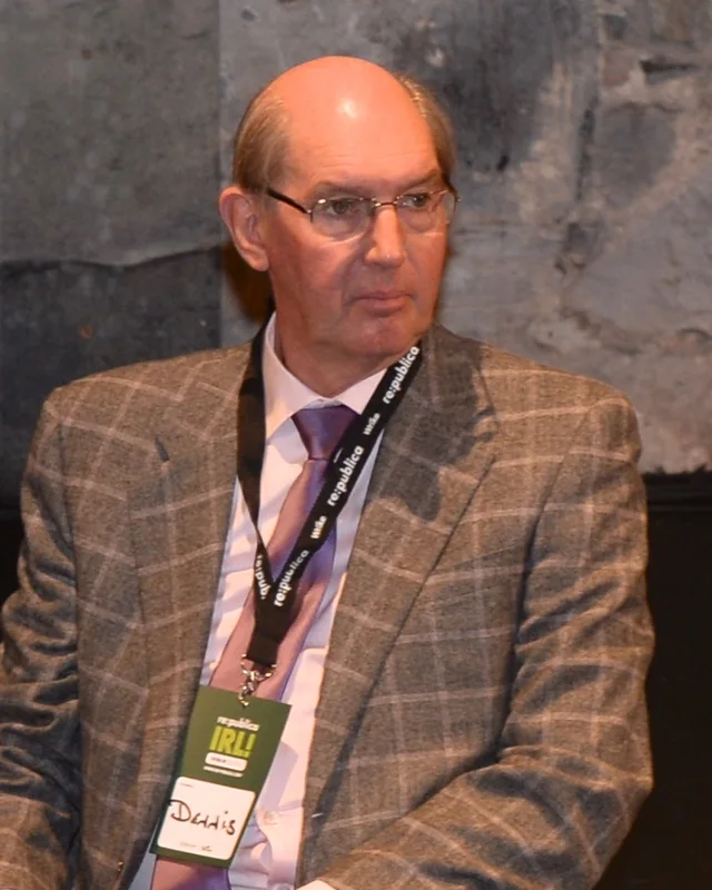 Dennis Jennings seated on a conference panel, in a checked jacket and tie, wearing a lanyard and a name badge reading Dennis.