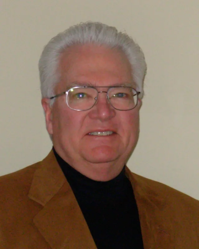 Douglas Gale in a studio headshot, in a tan jacket over a black rollneck, wearing wire-rimmed glasses.