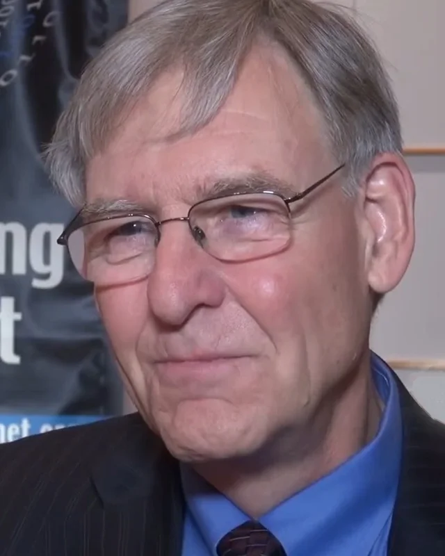 Doug Van Houweling in close-up during a recorded interview, in a dark suit and blue shirt, an Imagining the Internet banner behind him.