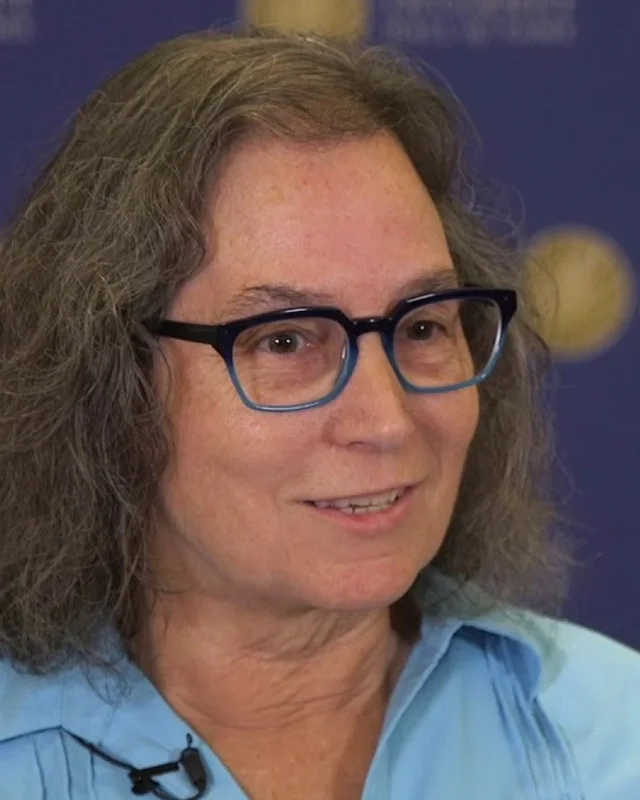 Elise Gerich in close-up during a recorded interview, in a blue shirt and dark-framed glasses, an Internet Hall of Fame backdrop behind her.