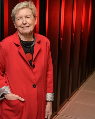 Jane Caviness standing beside a rack of network cabling in a machine room, in a red coat.