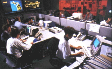 Color photograph of the Merit Network Operations Center: a dozen staff at desks of terminals in a low-lit room, with large wall displays showing a map of Michigan and a map of the national backbone, and racks of equipment lit red behind them.