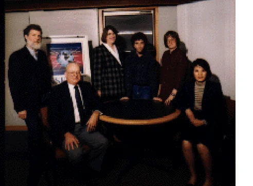 Six members of the Merit team in an office, gathered around a round table: five standing and one seated.