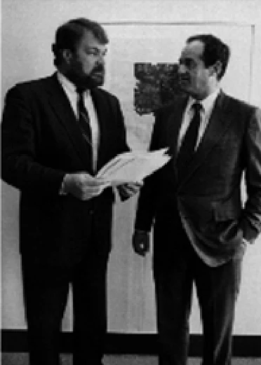 Two men in suits standing in an office, one holding a sheaf of papers and reading from it, the other looking on.